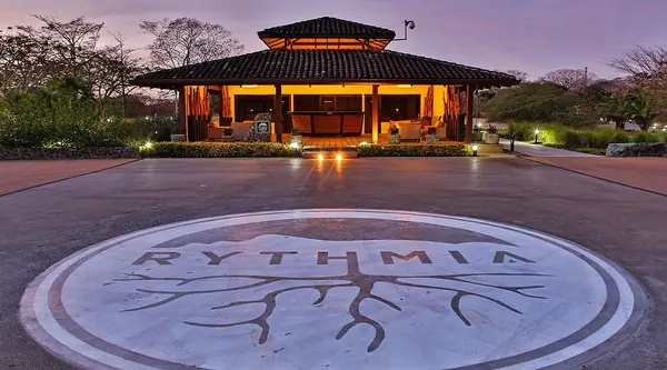 An open sided pavilion lit up at dusk, with the Rythmia logo set into the paving in front of it