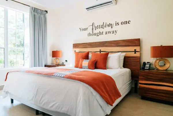 A guest bedroom with a large bed, orange cushions and throw, a wooden headboard and bedside tables, and the words "Your freedom is one thought away" on the wall