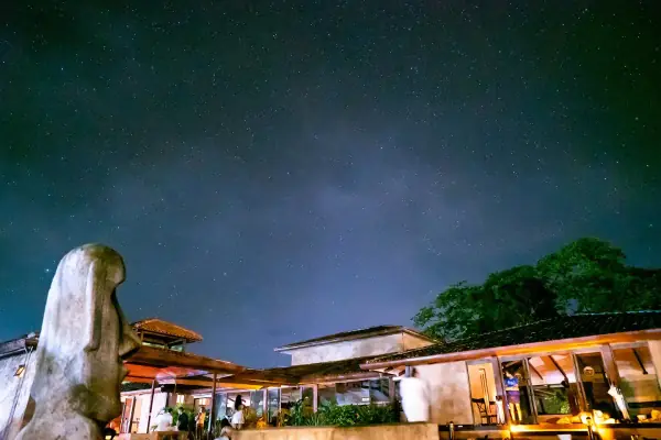 Rythmia's buildings at night under a sky full of stars, with a large carved stone head on the left and a few people outside